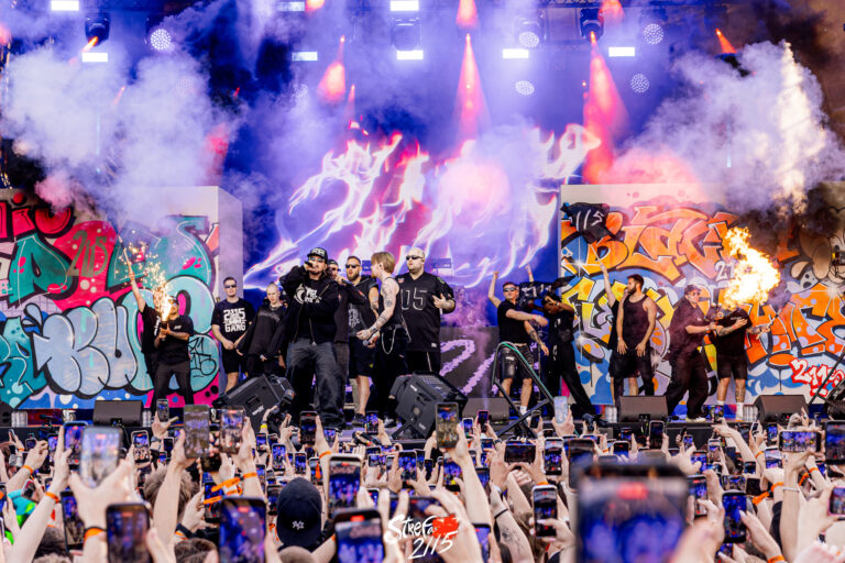 Grupa hip-hopowych wykonawców i tancerzy na scenie przed kolorowym graffiti STREFA, otoczona dymem, efektami ognia i jasnymi światłami, podczas gdy duży tłum nagrywa koncert swoimi telefonami.