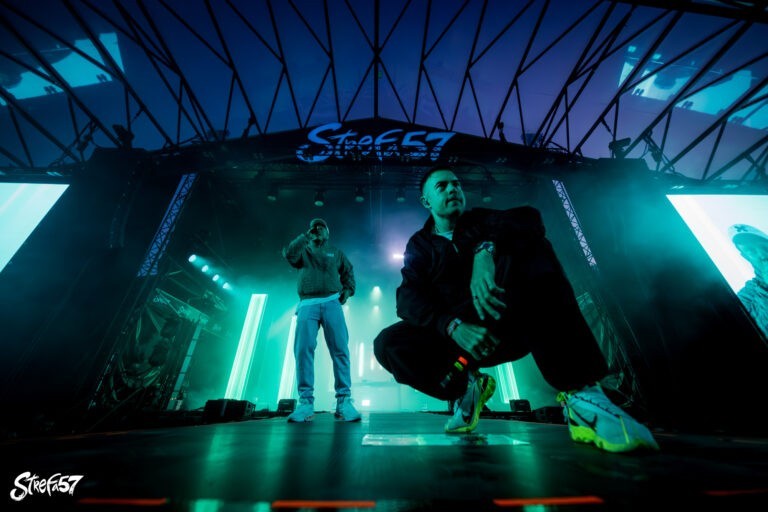 Dwóch wykonawców na scenie podczas nocnego wydarzenia muzycznego na świeżym powietrzu o nazwie STREFA 57; jeden stoi z mikrofonem, drugi przykuca na pierwszym planie. Scena jarzy się zielonymi i niebieskimi światłami, z dużym znakiem festiwalu i PRO8L3M nad głową.